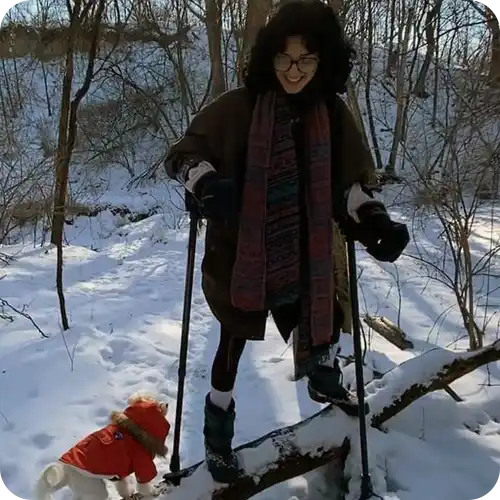 Person bundled up in winter clothing walking with ski poles in snowy woods.