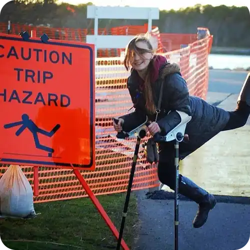 Person playfully posing next to a ’Caution Trip Hazard’ sign while using crutches.